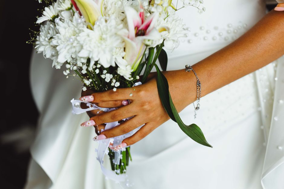 A close-up of a bride's hand holding a floral bouquet arranged with white lilies, white chrysanthemums, and delicate baby's breath. The bouquet is wrapped in white ribbon with small decorative accents