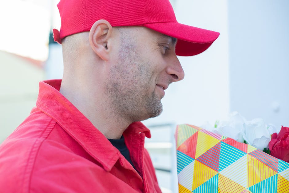 A delivery person dressed in a bright red cap and matching red jacket holding a colorful gift box wrapped in geometric patterned paper with shades of yellow, blue, and red, containing a bouquet of whi