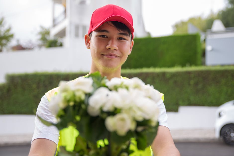 A delivery person wearing a red cap and a yellow reflective vest is smiling at the camera while handing over a bouquet of fresh white flowers, including roses and lilies, wrapped in clear cellophane. 